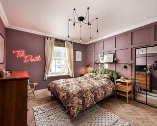 Hot pink neon light sign on aubergine hued wall, with dark floral bedlinen, wall paneling, and multi exposed bulbs ceiling pendant.