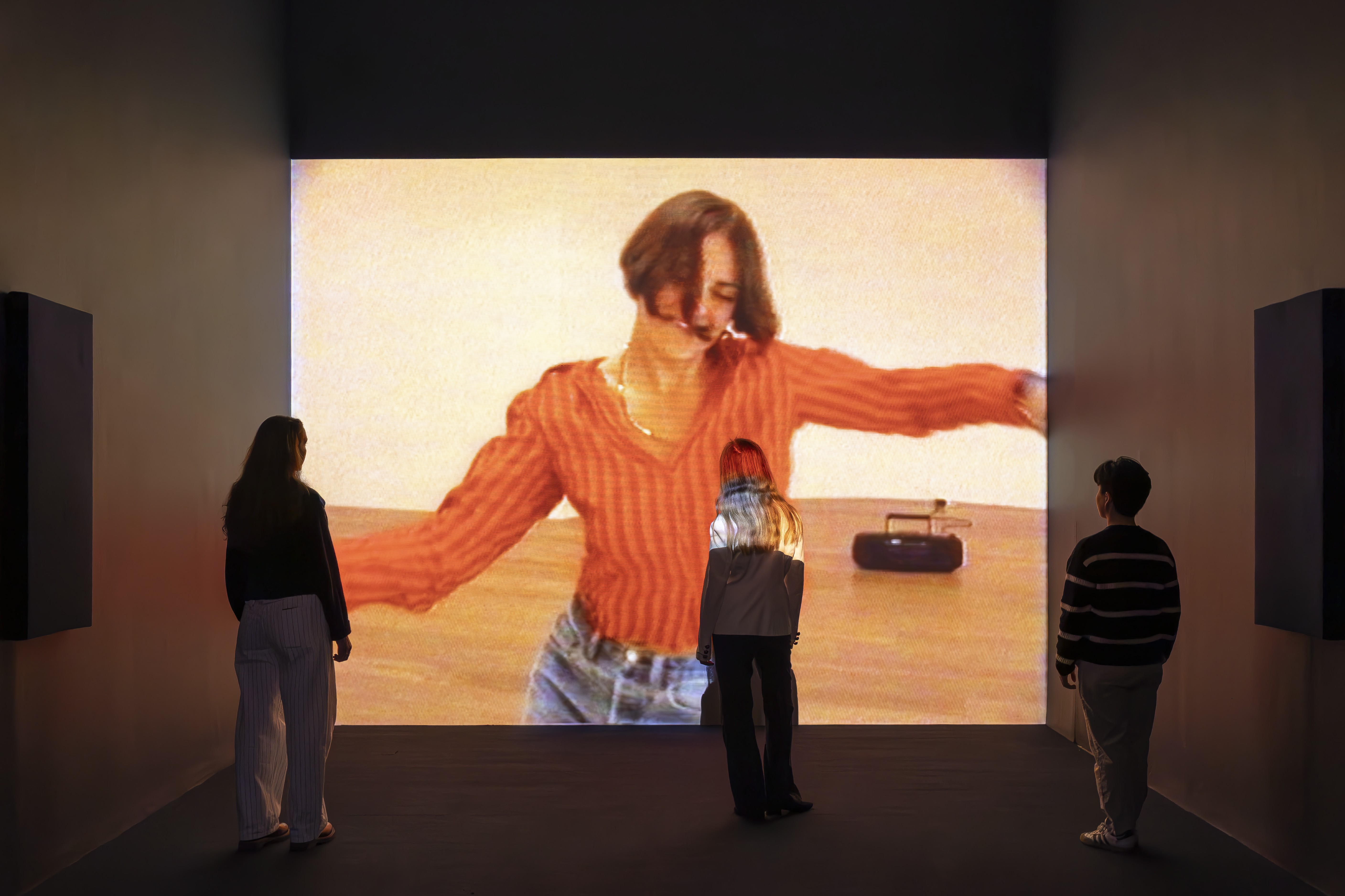 Three people stand in a gallery as a projection of a woman dancing, with short straight hair, a striped orange and red top, and denim jeans, plays on a wall before them.
