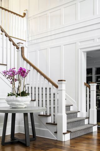 entrance way with table with large bowl of orchids, white painted panelling, gray staircase carpet, hardwood floor