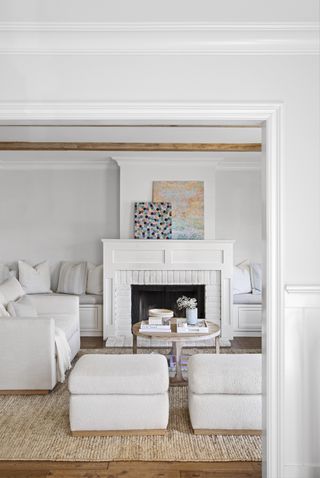 All white smooth couches and armchairs on a sand colored jute rug surrounding a light wooden square coffee table and facing two colorful canvases a top the panelled with and black mantel