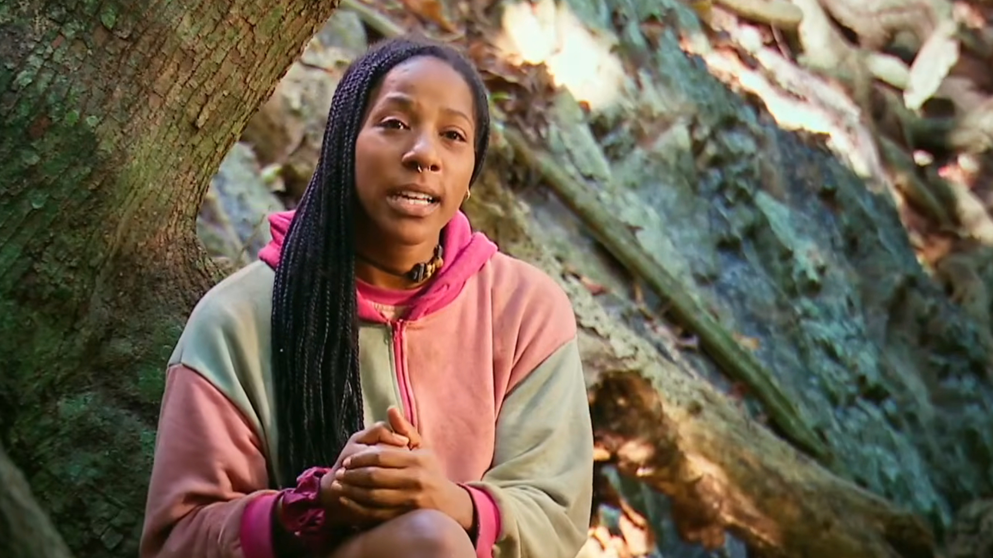 Tiffany talks while wearing a multi-colored sweater in front of a tree on Survivor.