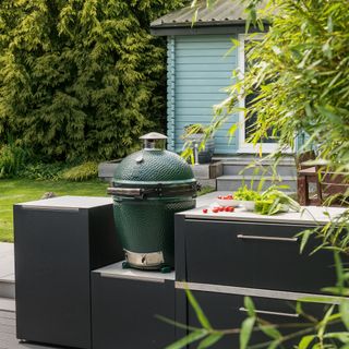 a compact outdoor kitchen with several units and a big green egg BBQ, with a duck egg blue painted wooden shed in the background