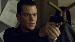 A close-up of Matt Damon playing Jason Bourne in The Bourne Trilogy of movies while holding a gun and wearing a black top.