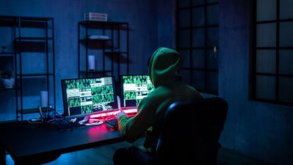 A hooded computer operator in a darkened room