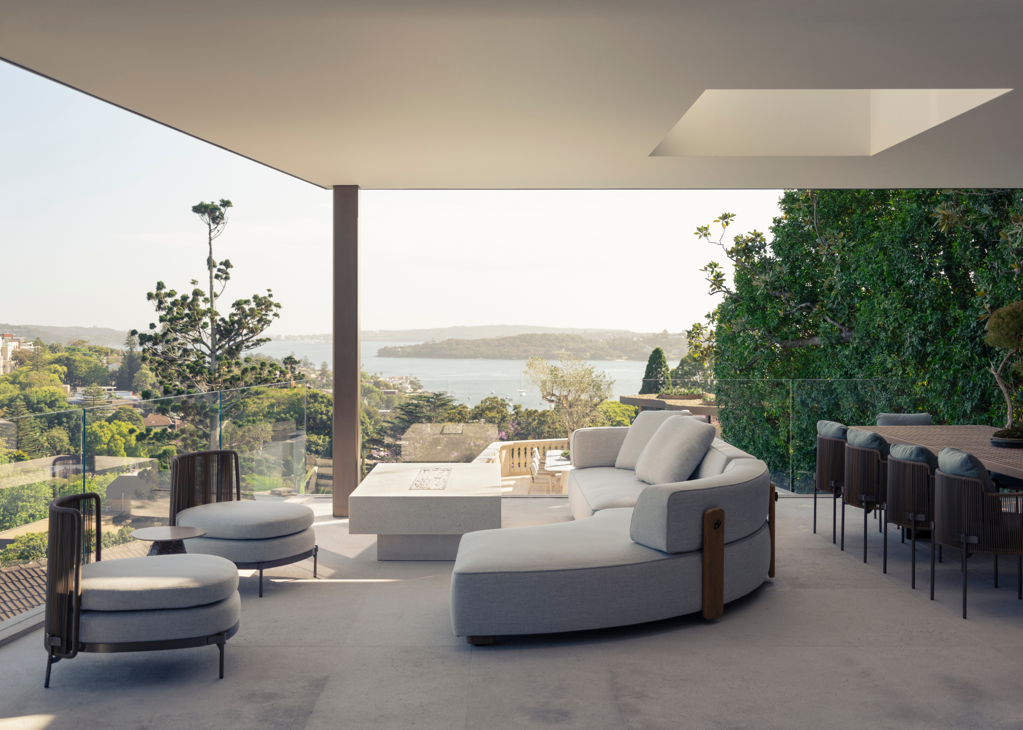 An outdoor living room with a pair of gray chairs, a firepit table and a curved couch by a dining space