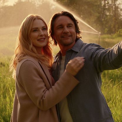 alexandra breckenridge as melida and martin henderson as jack smiling and looking out into a field while a sprinkler goes off behind them in a still from virgin river season 7