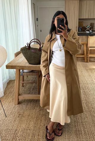 a photo of a summer rainy day outfit with a woman wearing a tan trench coat over a white t-shirt and off-white satin slip skirt with brown flip flops and a woven tote bag