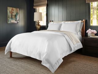 White bed sheets against dark olive green tongue and groove style slat walls in a bedroom. Abstract art sits opposite two windows and striped Roman blinds.