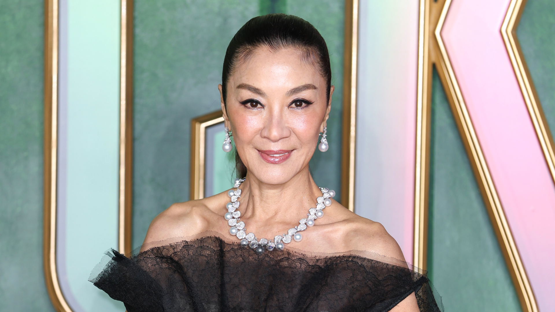Michelle Yeoh wearing a ponytail hairstyle. She is also wearing a black off the shoulder dress, a diamond necklace, and earrings. 