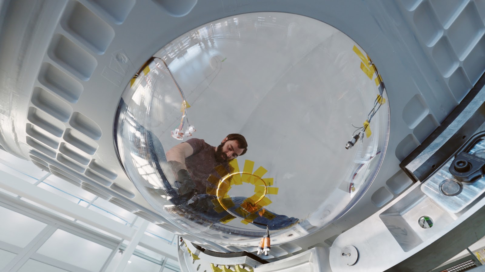 A man stands above a domed pane of glass with a wire in secured with yellow tape at its center in the shape of a spiral. The dome is secured by metal sheets of a space module.