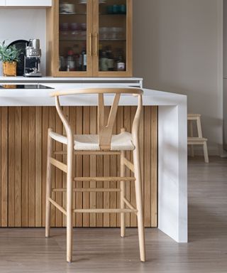 A white kitchen island with a waterfall edge, a wooden base, a curved wooden chair in front of it, and a countertop with a coffee maker and wooden cabinet on top