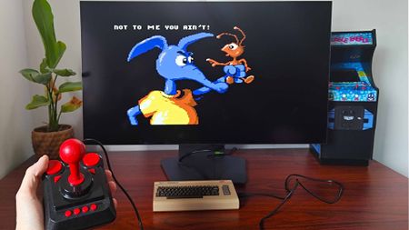 Hand holding C64 Mini joystick with console connector to monitor in front with Nobby the Aardvark gameplay on screen.