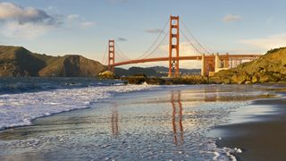 The gold gate bridge in San Francisco is reflected on a wet, sandy beach at sunset.