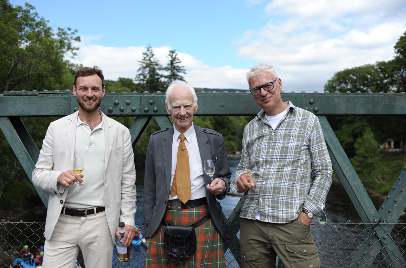 Three men stand on a bridge holding glasses of whisky