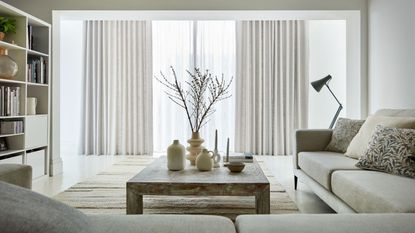 A modern white living room with neutral decor and sliding glass doors with sheer curtains and a heavier neutral curtain. 