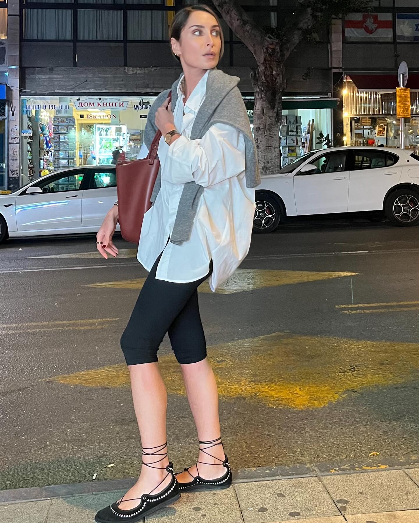 influencer Géraldine Boublil poses on a city sidewalk wearing an oversize white button-down shirt, a sweater draped over her shoulders, brown tote bag, black capri pants, and black lace-up flats