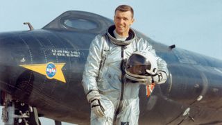 a smiling man in a silver flight suit stands in front of a black plane on a runway
