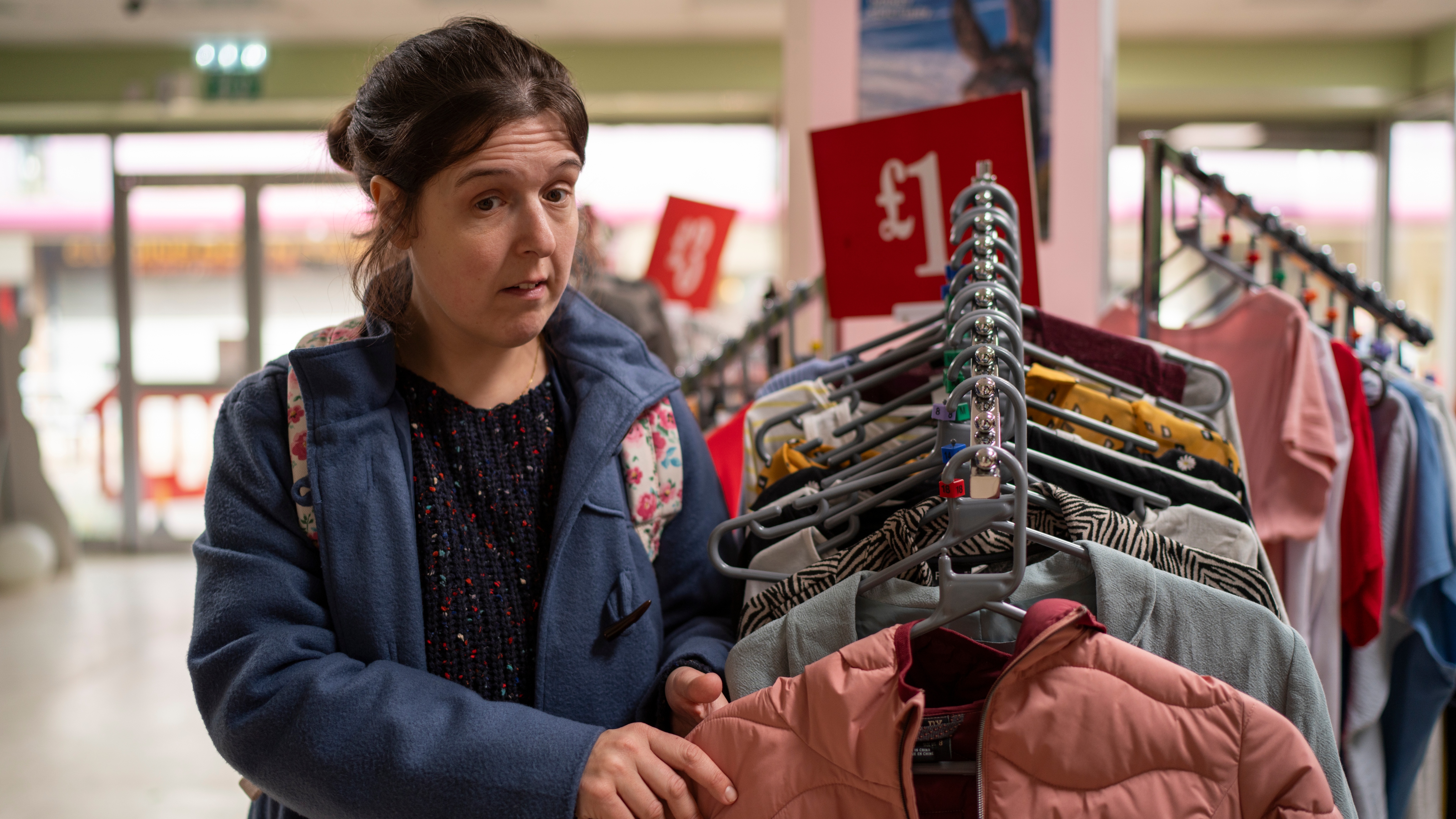 Rosie Jones as Emily standing by a rack of clothes in a shop.