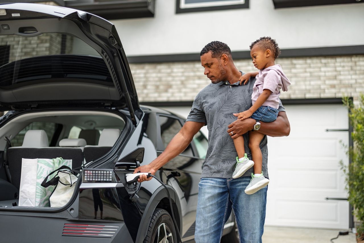 A man holds his young son while charging his electric car at home. The hatch is open and it&#039;s clear he has gone grocery shopping.