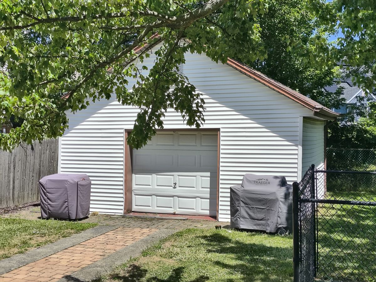 White wooden garage surrounded by trees and green grass with two outdoor chairs under weather-proof covers in the sun