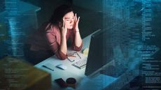 Female cybersecurity analyst pressing fingers to her temple in despair while working at a desktop computer in a dimly lit room.