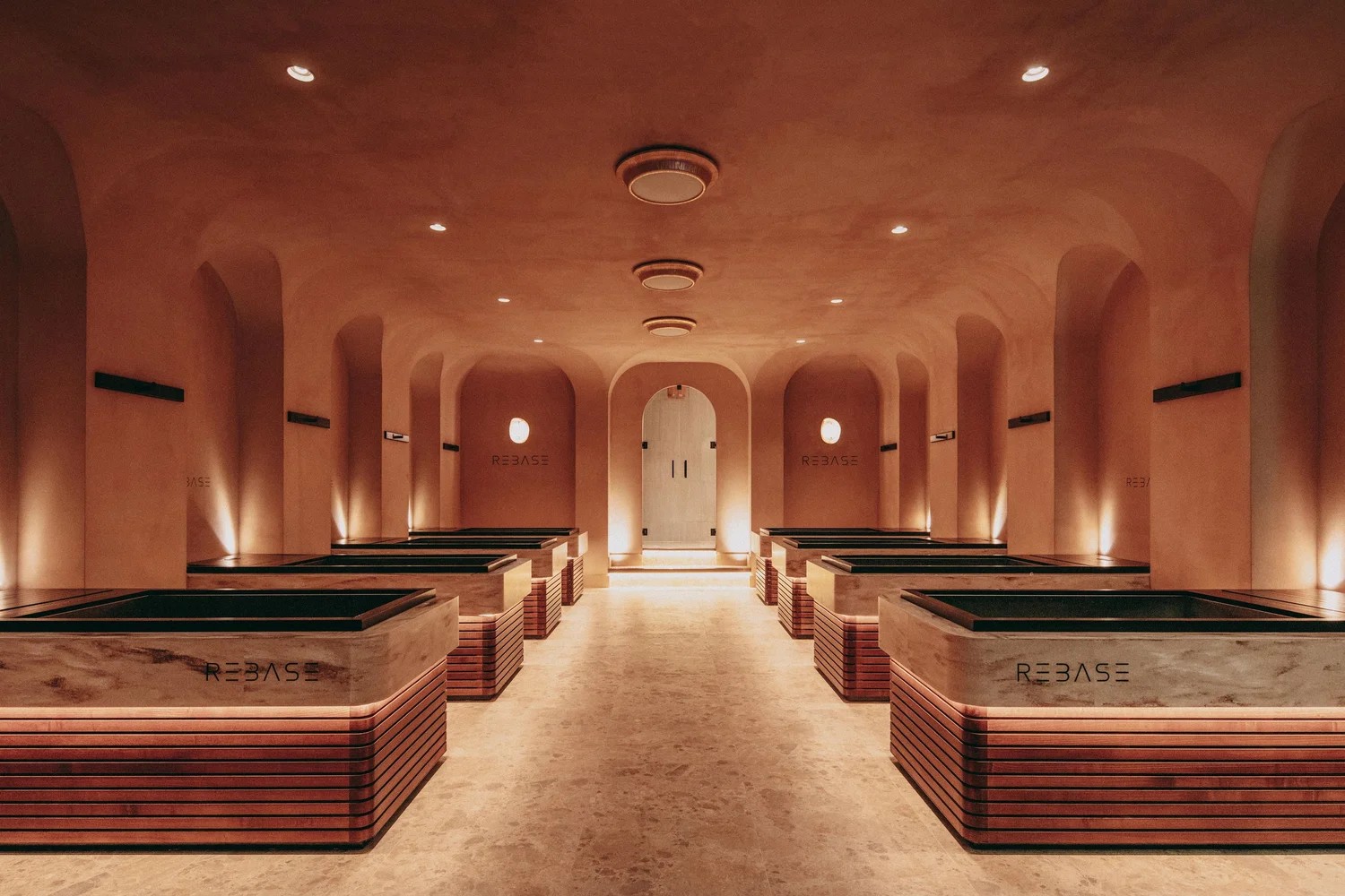 A warmly decorated, contemporary sauna hub with creamy and terracotta plaster walls, curvilinear upholstered furniture with green fabric, wooden details, and warm pendant as well as spherical sconce lighting throughout.