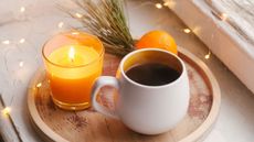 A wooden circular serving platter holding a lit candle, a white mug with coffee, and an orange on a white windowsill with fairy lights scattered about