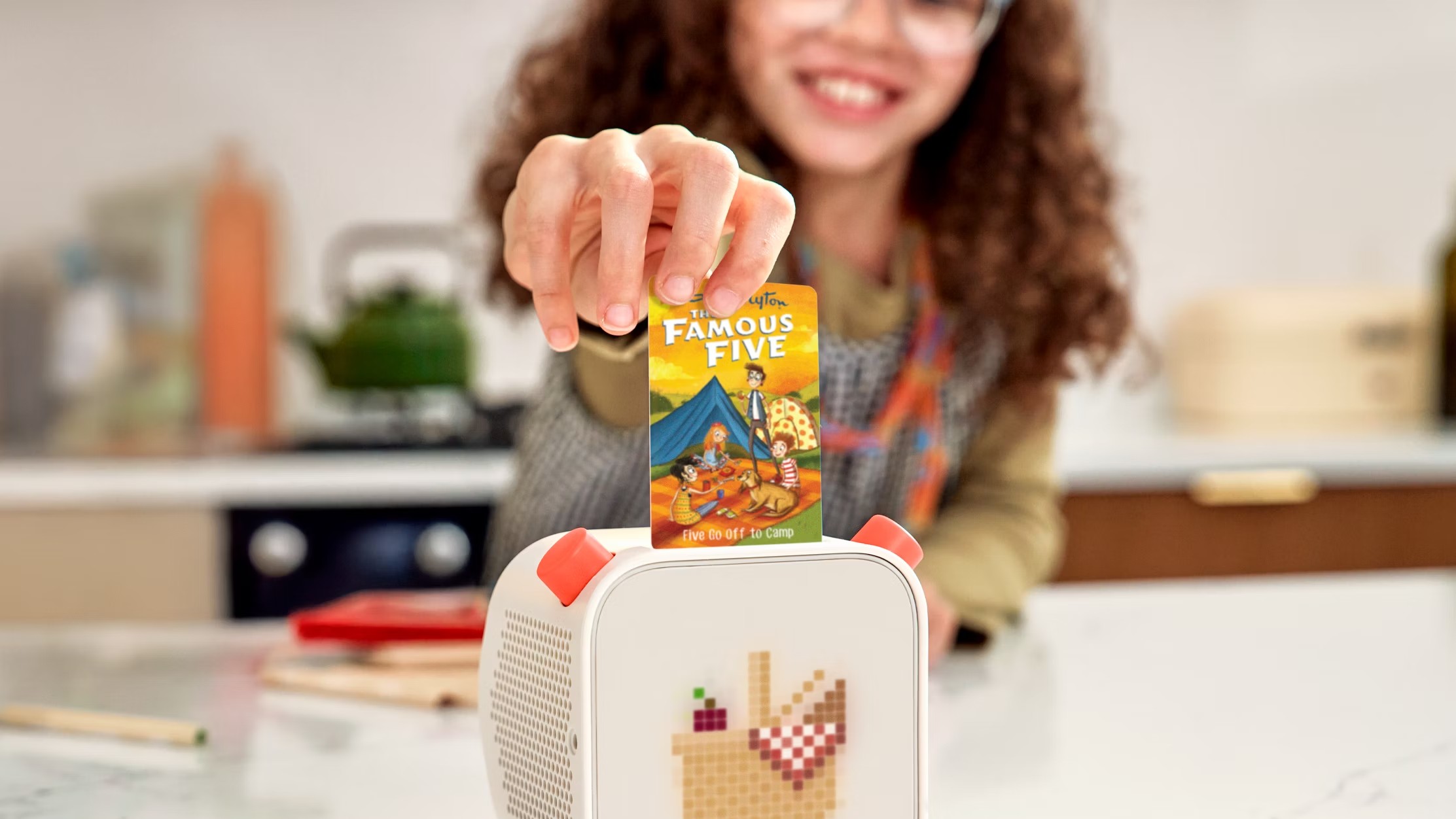 A child, smiling and wearing glasses, inserts a bright orange "The Famous Five: Five Go Off To Camp" story card into a white, cube-shaped digital audio player with a pixelated picnic basket display.
