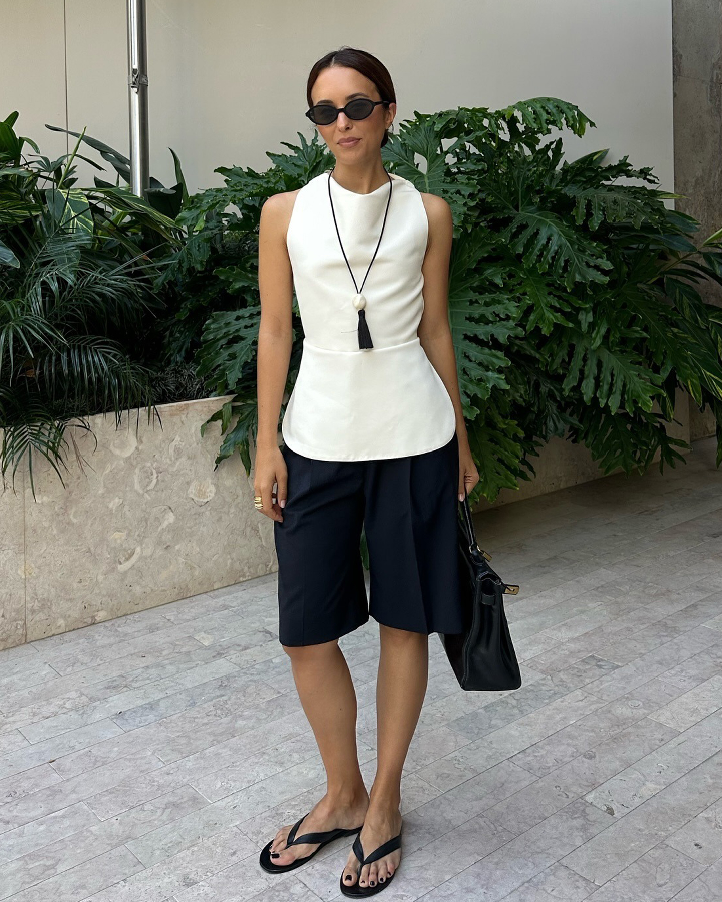 Style influencer Debora Rosa poses in Lisbon, Portugal wearing black sunglasses, black cord necklace with pendant, draped sleeveless white top, black Bermuda shorts, and black flat flip-fop sandals.