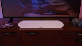 A white Sonos Ray soundbar on a TV cabinet beneath a flatscreen TV in dim lighting.
