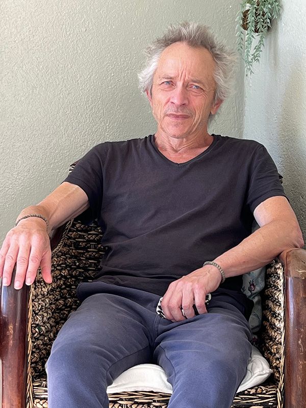 A man with short white hair wearing a black t-shirt and blue jeans sits in a brown, woven chair, looking at the camera