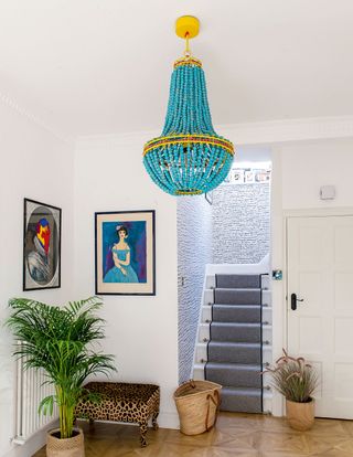 A white hallway with framed wall art, leopard print stool, statement blue and yellow chandelier ceiling light, white staircase with grey staircase runner and grey printed hallway wallpaper