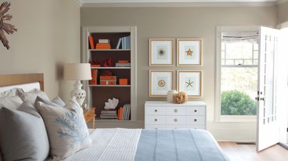 Coastal bedroom with Pashmina by Benjamin Moore walls, a bed with pale blue and white bedding, a wall of nautical prints, a white dresser, and built-in bookshelves with orange boxes.