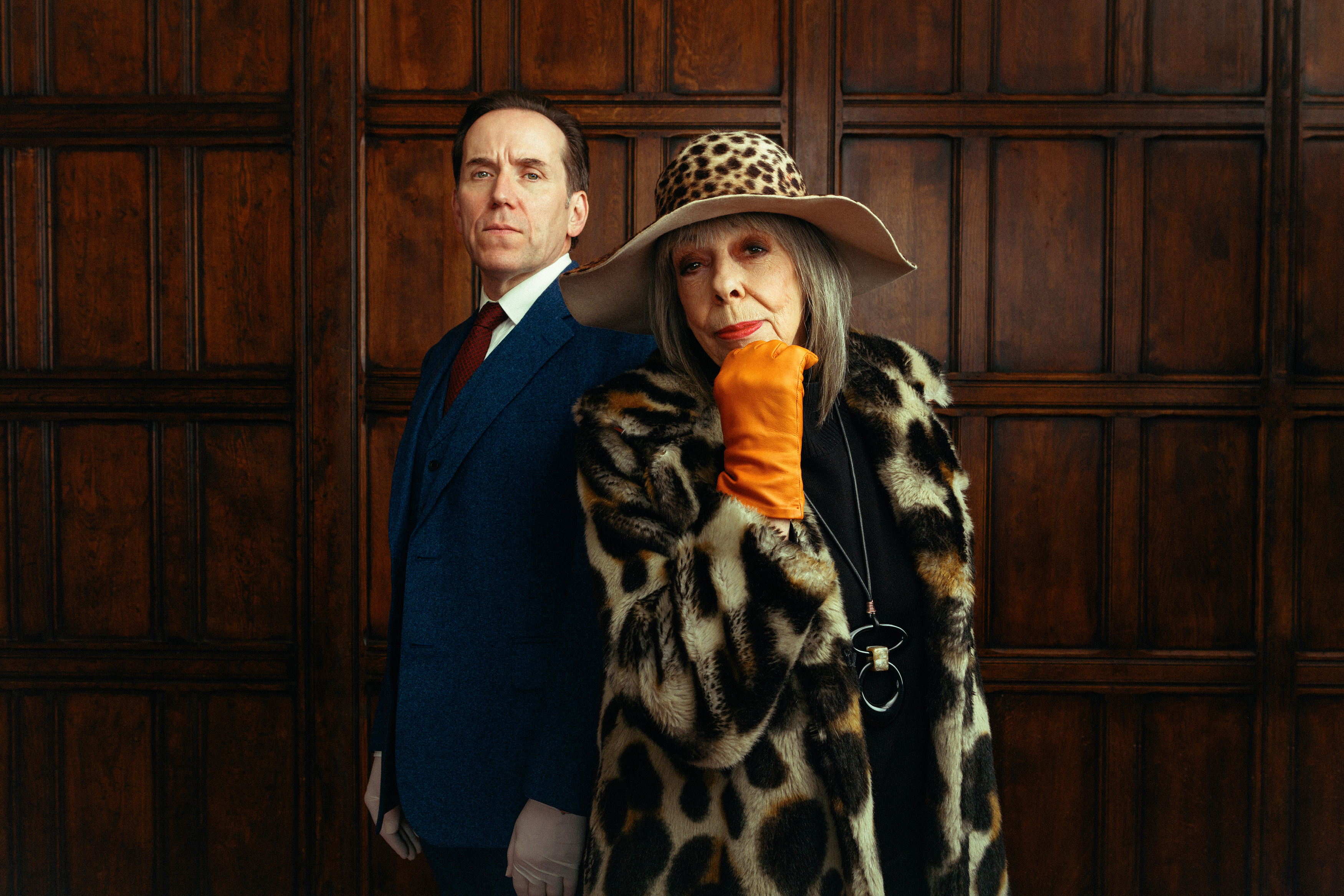 Professor Jasper Tempest (Ben Miller) and his mother Adelaide (Frances de la Tour) stand in front of a mahogany panelled wall. He is wearing a suit and white plastic gloves, and standing upright. She is wearing a large fur coat and a leopard-print wide-brimmed hat accessorised with orange leather gloves, and is leaning forward with her chin resting on her right hand.