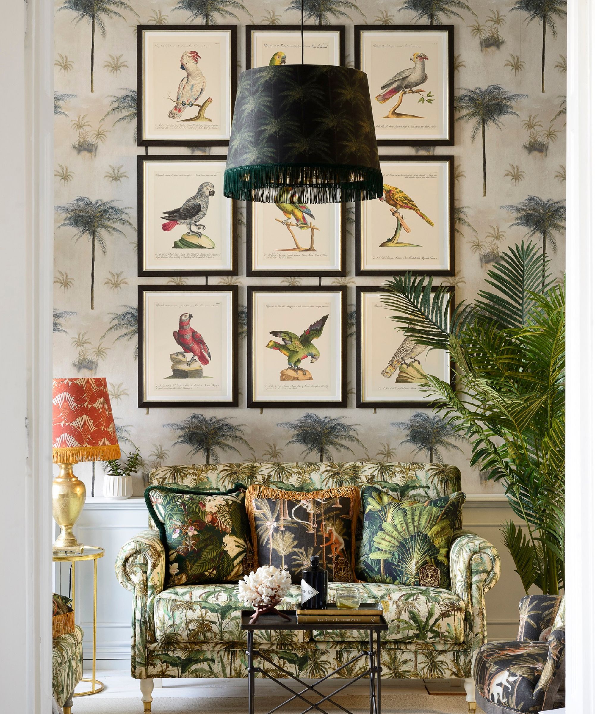 A living room with tropical wallpaper covered in palm trees. A sofa with green patterned fabric sits below framed bird illustrations. Cushions, plants and a fringed lampshade add colour, softness and an exotic, layered feel to the relaxed interior space.