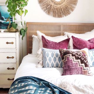 Colorful boho inspired bed with cane headboard, natural texture wall piece, trailing houseplant, and assorted patterned and block printed scatter pillows in plum and blue hues.