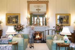 Queen Elizabeth wearing a sweater and skirt standing in front of a fireplace and holding a cane at Balmoral Castle