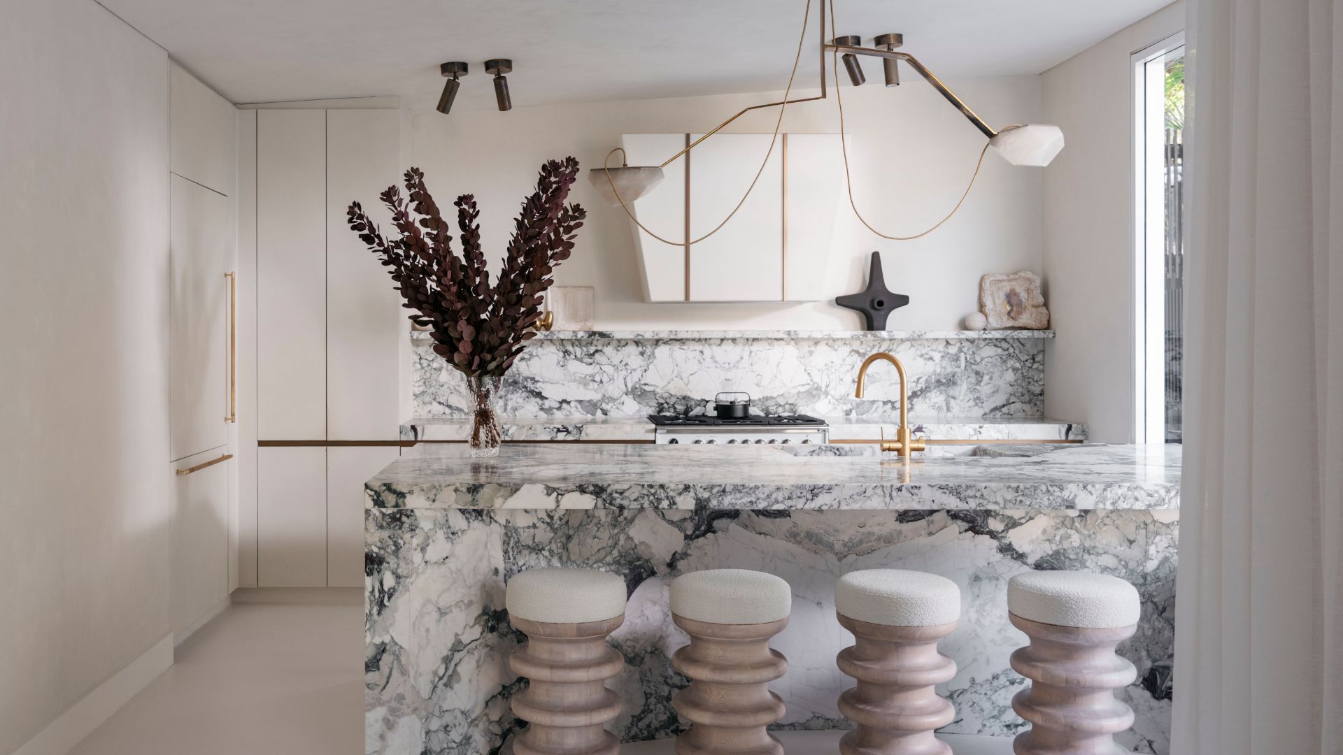 A stunning kitchen with a marble island and backsplash, a set of four sculptural stools, a vase of dried stems, and storage cupboards with gold hardware
