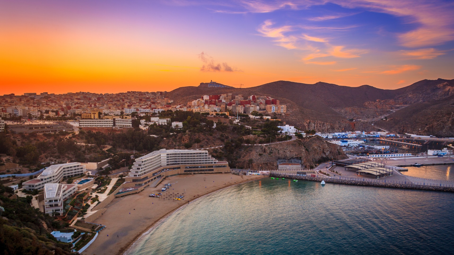 Aerial view of a coastal town and crescent beach at sunset, with hillside buildings, calm water, and vivid orange-pink skies creating a serene mood.