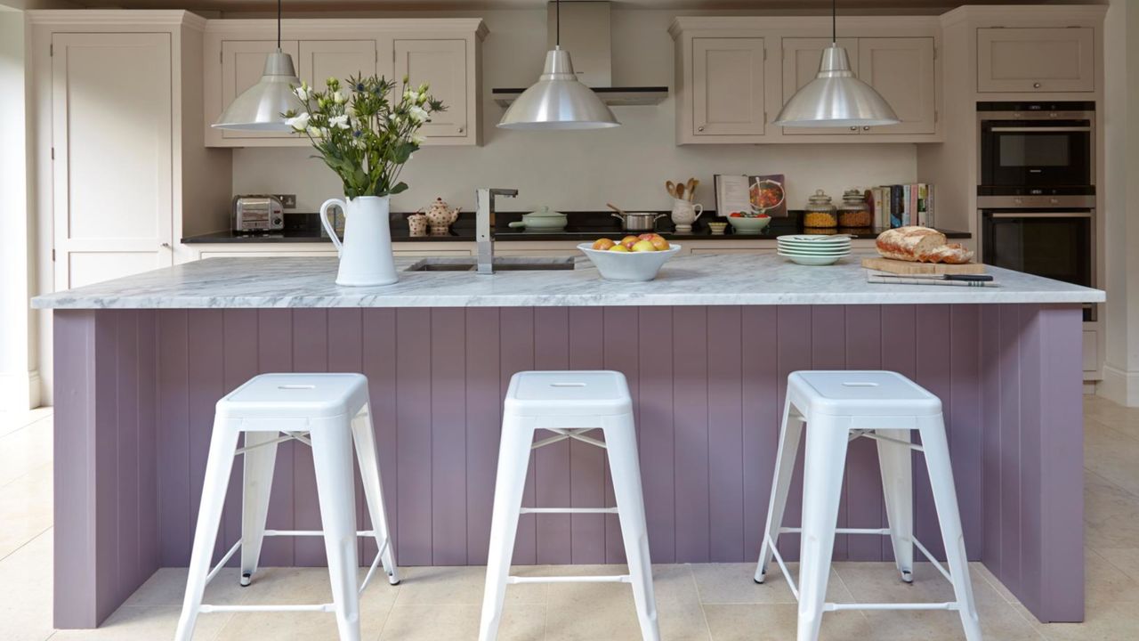 lilac kitchen island with white and grey worktop, cream cabinets, white stools and grey pendant lights