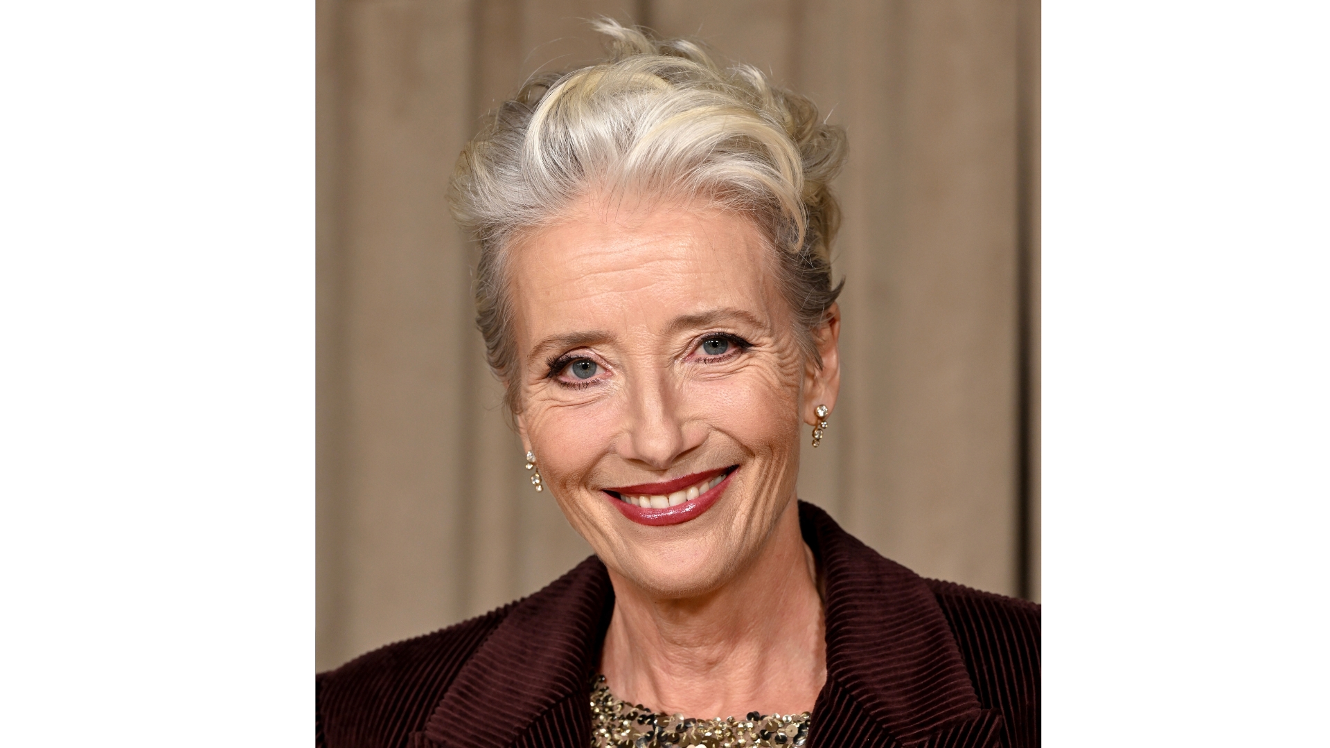 Image of Emma Thompson smiling with a grey bouncy cropped haircut, wearing a burgundy blazer and deep red lipstick