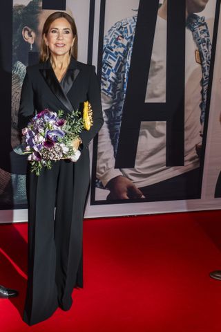 Queen Mary wearing a black jumpsuit standing on the red carpet and holding flowers