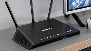 Lifestyle image of the Netgear Nighthawk R6700 Wi-Fi Router on a desk next to a computer monitor.