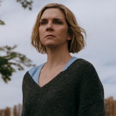 rhea seehorn as carol wearing a grey sweater standing in her yard and staring into the distance in pluribus season 1