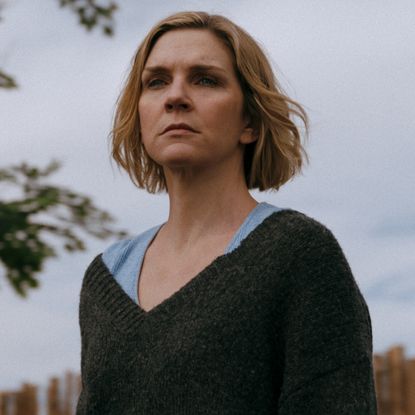 rhea seehorn as carol wearing a grey sweater standing in her yard and staring into the distance in pluribus season 1