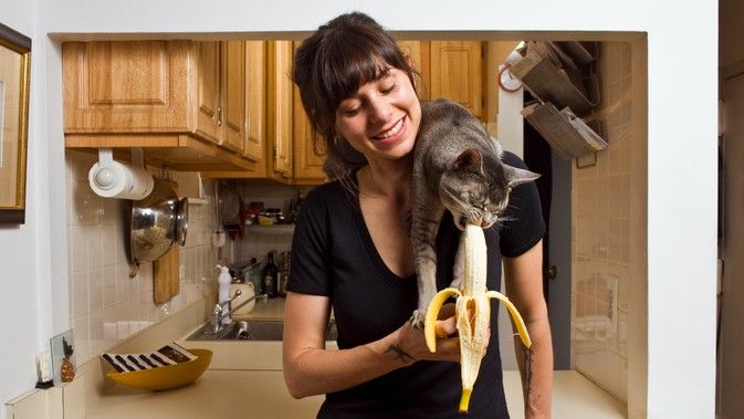 A woman feeding her cat a banana, the cat is sat on her shoulder