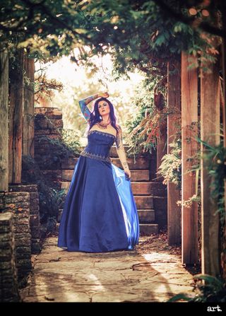 An image of a woman with purple hair in a blue ballgown, standing in paved garden with a canopy of leaves, taken with the Olympus Vintage I Art FIlter