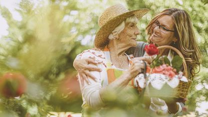 An older woman carrying a small basket of greenery and her daughter embrace in the garden.