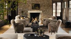 a moody modern farmhouse living room with a large stone fireplace and four sheepskin armchairs styled by the fire on a large rug with dark black wooden rustic tables
