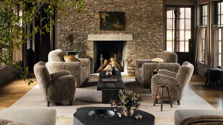 a moody modern farmhouse living room with a large stone fireplace and four sheepskin armchairs styled by the fire on a large rug with dark black wooden rustic tables